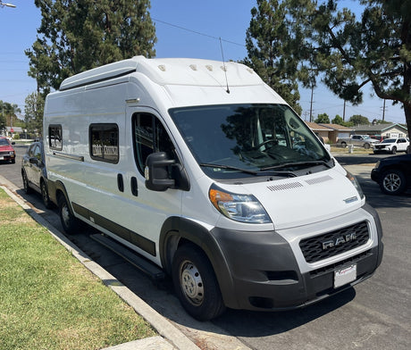 Bright white Ram ProMaster camper van, an rv rental, parked curbside on a sunny residential street. Equipped with windows & roof AC.