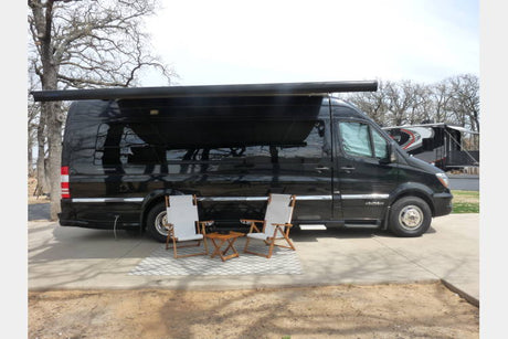 Black Sprinter RV with extended awning, two chairs, and table at a campsite. Ideal for comfortable RV rental travel and outdoor leisure.