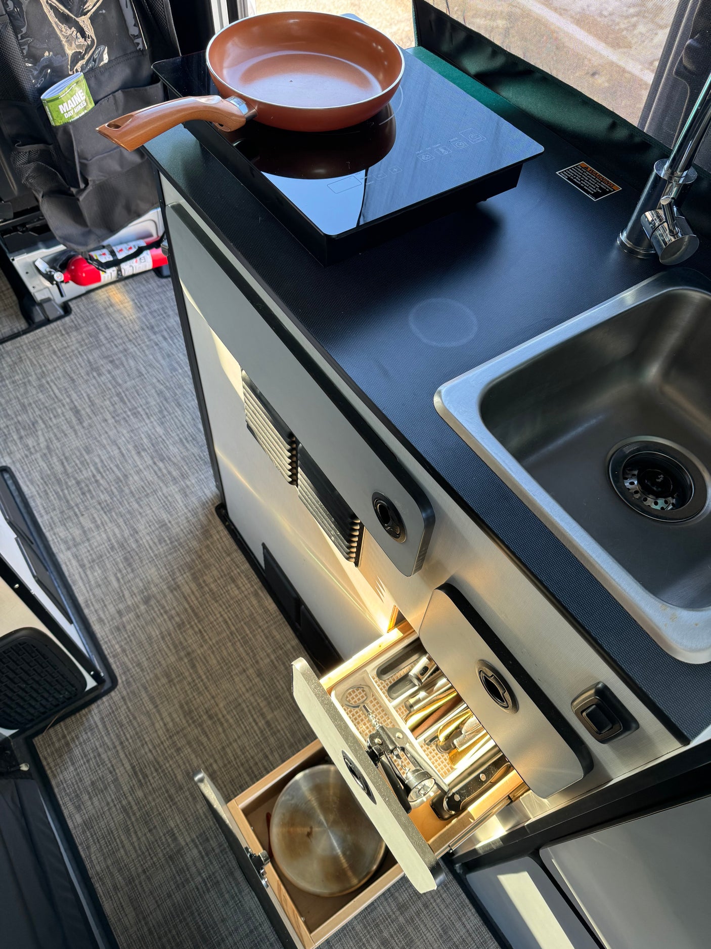 Explore this RV rental's compact kitchen: induction cooktop, sink, & organized drawers with utensils & pots. Fire extinguisher visible.