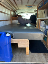 Cozy RV rental camper van interior: gray bed, custom wood shelving, a blue water jug, and ceiling fan. Ready for travel.
