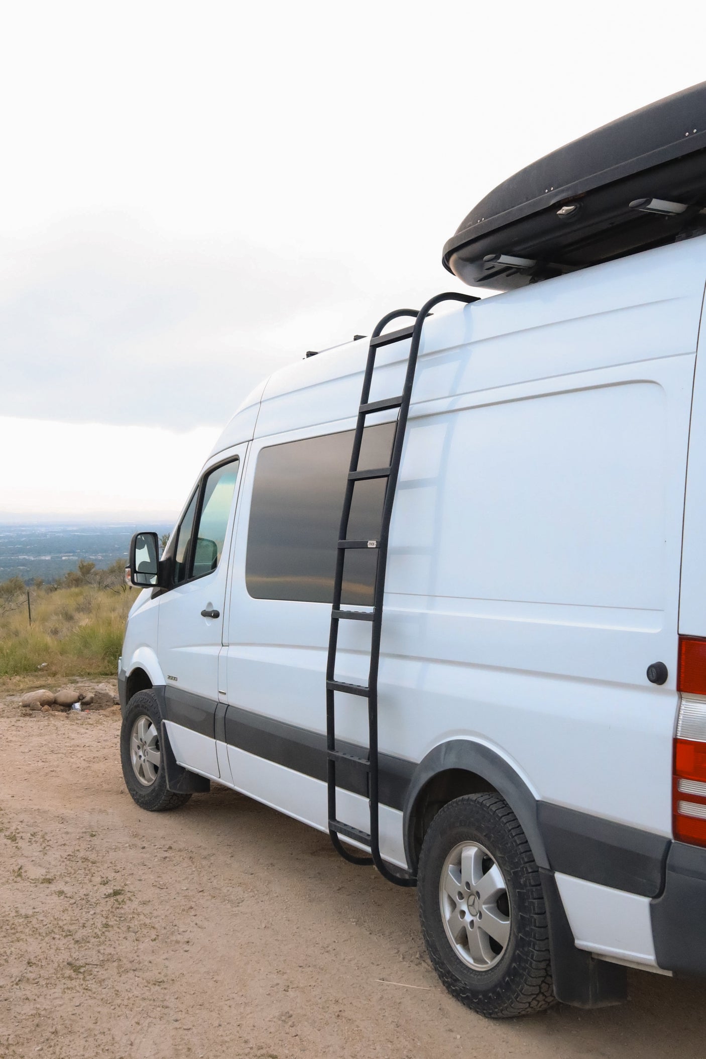 White camper van with a roof cargo box & ladder, on a dirt road with a scenic view. Perfect for an RV rental adventure!