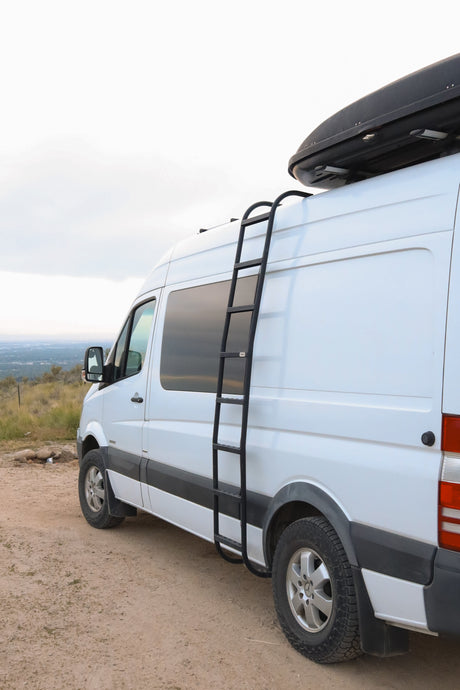 White camper van with a roof cargo box & ladder, on a dirt road with a scenic view. Perfect for an RV rental adventure!