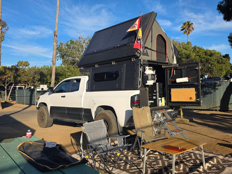Overland truck with rooftop tent set up at an RV rental site. Camping chairs & table await for a relaxing adventure.|Explore off-road with this RV rental: a white Toyota Tundra truck with a rooftop tent