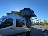 Silver RV rental camper van with a deployed rooftop tent and an extending ladder, parked near a Holiday Inn.