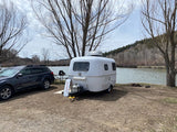 An rv rental, a white travel trailer, and an SUV parked at a serene lakeside campsite with bare trees and a mountain.