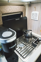 RV rental kitchen featuring a Keurig, stovetop with kettle, and a "Bacon Is My Love Language" sign. Compact and functional for travel.