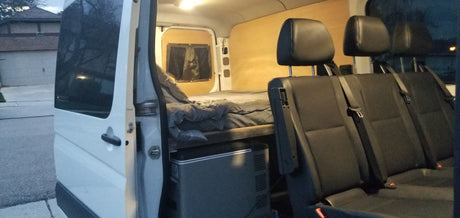 This inviting RV rental van's interior features a cozy bed, portable cooler, and three passenger seats, visible from the open side door.