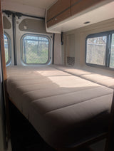 Bright rv rental interior with two beds, overhead cabinet, and side windows revealing trees and a fence outside, bathed in sunlight.