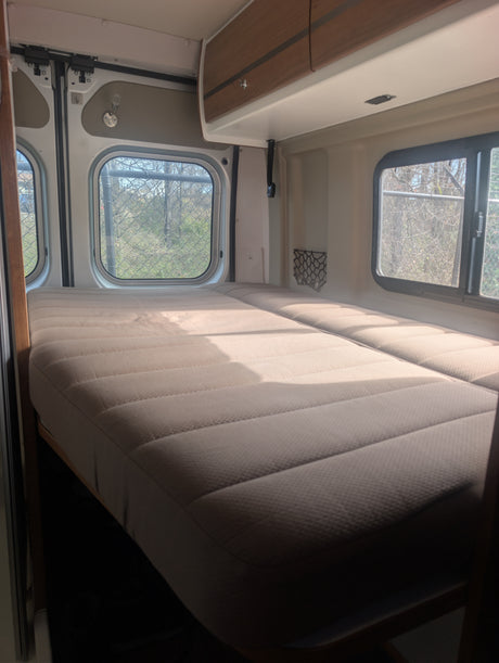 Bright rv rental interior with two beds, overhead cabinet, and side windows revealing trees and a fence outside, bathed in sunlight.