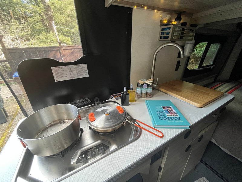 RV rental interior showing a cozy kitchen with a Suburban stovetop, pots, and a "Van Life Cookbook" on the counter. — photo 9