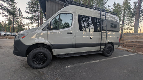 Adventure-ready white Mercedes Sprinter RV rental with off-road tires and roof rack, parked in a scenic forest setting.