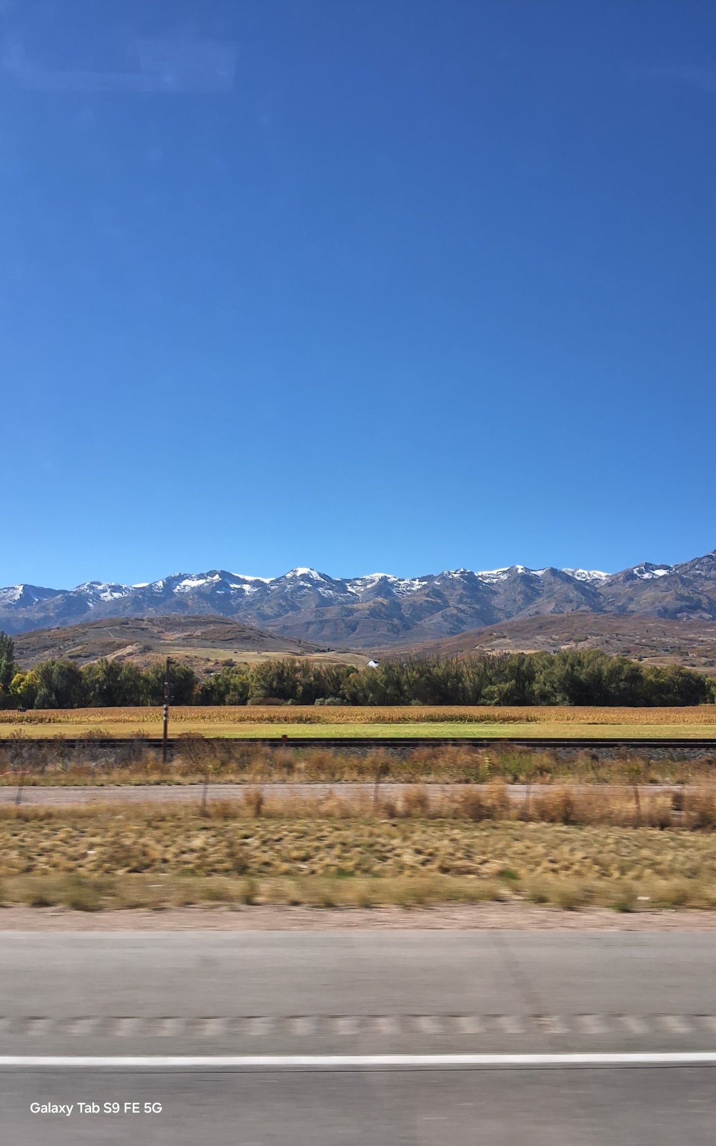 Scenic snow-capped mountains, golden fields, and clear blue sky from a moving vehicle. Perfect for an RV rental road trip.