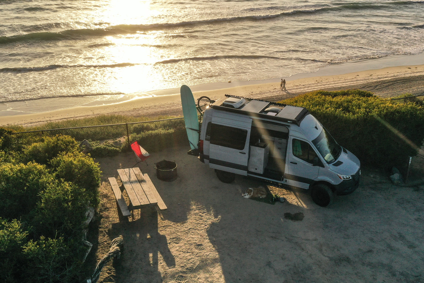 Beachfront RV rental setup: campervan, surfboard, dog, picnic, and fire pit at sunset with ocean waves.