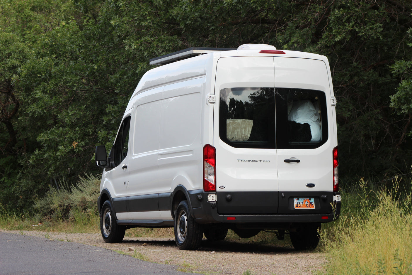 White Ford Transit 250 camper van with solar panels for RV rental, parked roadside by green trees.