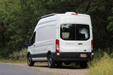 White Ford Transit 250 camper van with solar panels for RV rental, parked roadside by green trees.