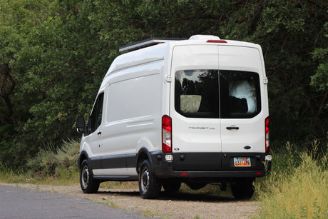 White Ford Transit 250 camper van with solar panels for RV rental, parked roadside by green trees.