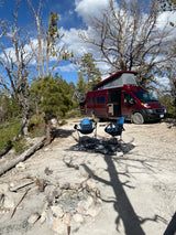 Scenic **rv rental** camping setup: Red van with bikes, chairs, & fire pit in a dry, forested wilderness area.