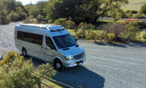 Silver Mercedes Sprinter RV rental, a sleek camper van, parked on a gravel road under sunny skies, surrounded by greenery.