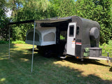 Compact RV rental: a dark grey travel trailer with an extended awning and pop-out tent, nestled in a lush green campsite.