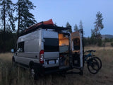 A silver camper van (RV rental) with open back doors, showing interior, kayak on roof, and bikes. Parked in a field at dusk.