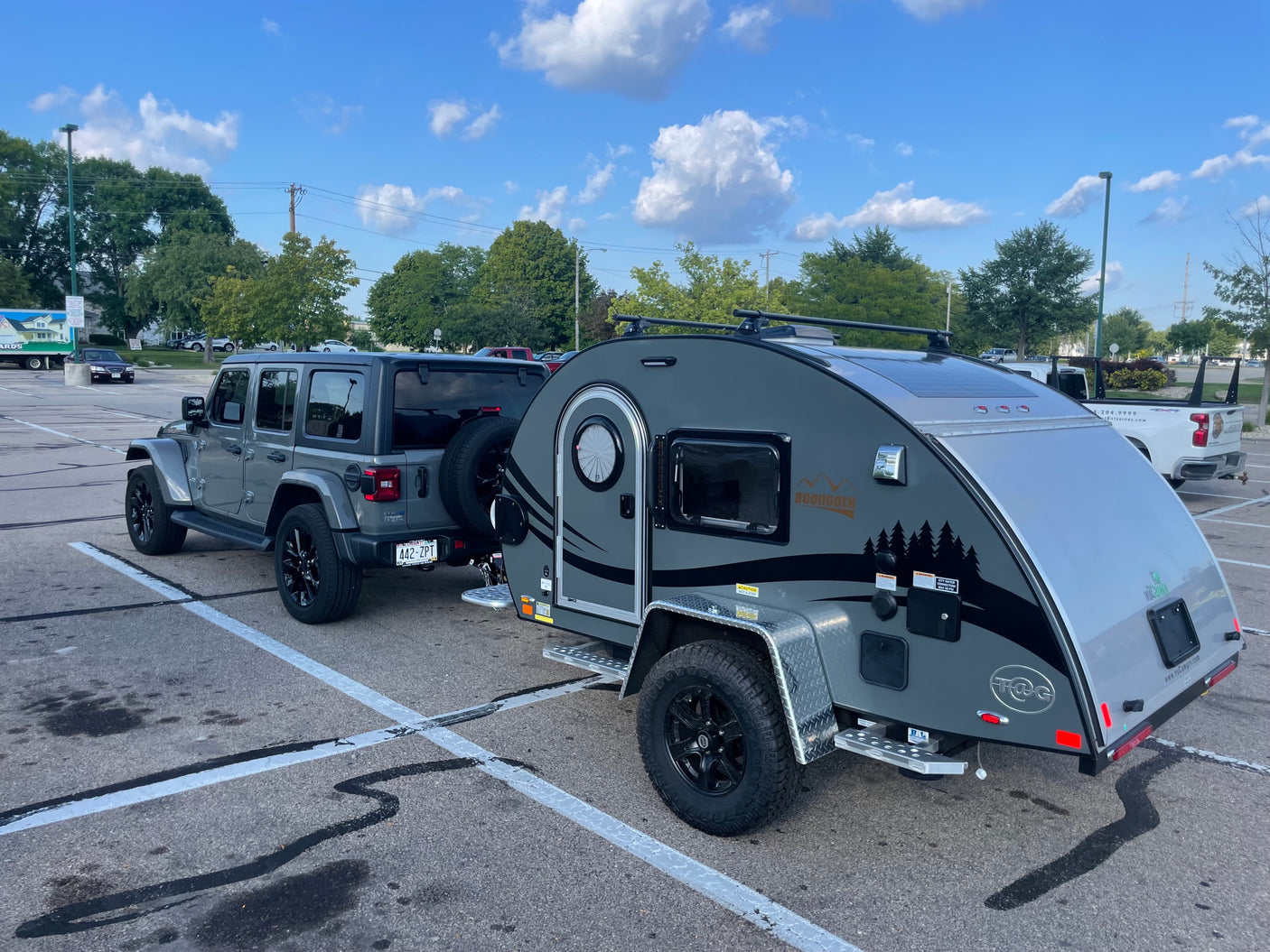 A grey Jeep Wrangler tows a sleek nuCamp Boondock teardrop RV camper in a parking lot, ready for an RV rental adventure.