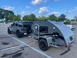 A grey Jeep Wrangler tows a sleek nuCamp Boondock teardrop RV camper in a parking lot, ready for an RV rental adventure.
