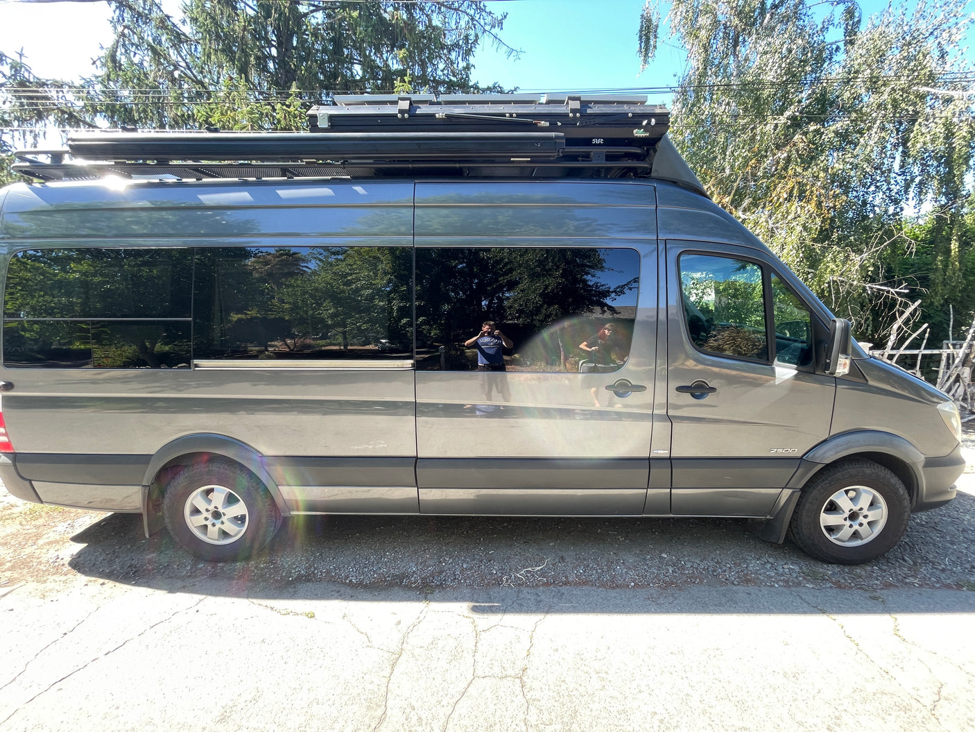 Grey Sprinter camper van, likely an RV rental, with a roof rack and tinted windows reflecting trees & people.
