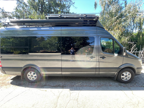 Grey Sprinter camper van, likely an RV rental, with a roof rack and tinted windows reflecting trees & people.