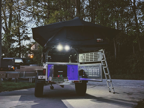 Off-road RV rental: a camping trailer with rooftop tent, ladder, and illuminated power/storage setup.