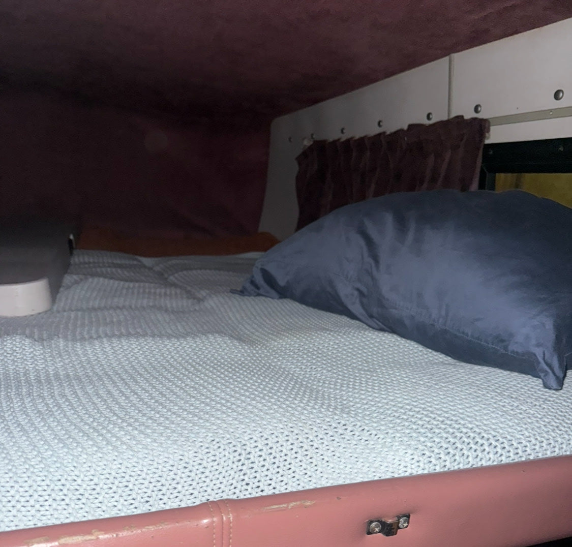 Cozy sleeping area in an RV rental, showing a bed with a blue pillow and light knitted blanket under a low ceiling.