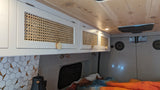 RV rental interior: Overhead white cabinets with rattan panels, wood ceiling, hexagonal backsplash, and a cozy bed area.