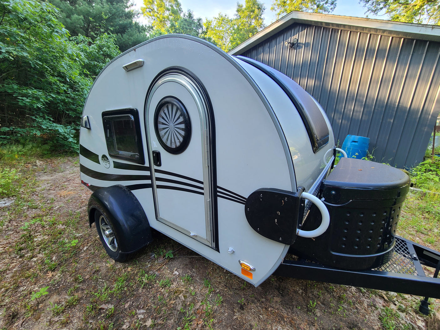 A white teardrop camper with black stripes, ideal for an RV rental, is parked near a dark gray metal building.