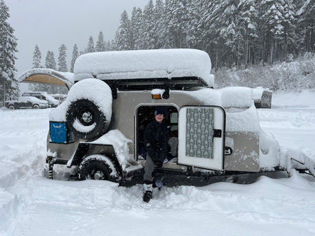 A smiling child in ski boots exits a small, snow-covered RV rental travel trailer amidst a very snowy forest.
