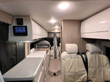 Inside an RV rental, a compact living space features a kitchen, bed area, passenger seats, and a wall control panel.