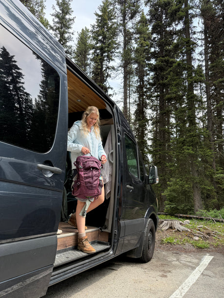 Woman with purple backpack exits her rv rental camper van in a forest, ready for outdoor adventure.