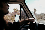 Driver's view from an RV rental on a desert road trip, featuring Joshua trees and rocky hills through the windshield.