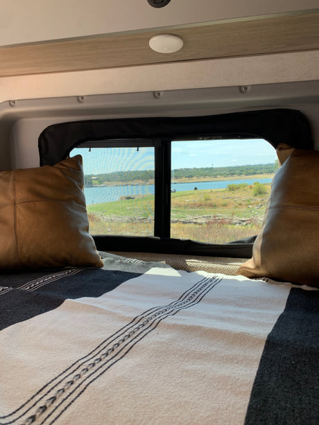 Comfortable RV rental bed with a scenic window view of a serene lake and natural landscape, ideal for travelers.