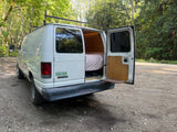 White RV rental camper van with open back, revealing a cozy bed and fold-down table, parked on a gravel road surrounded by trees.