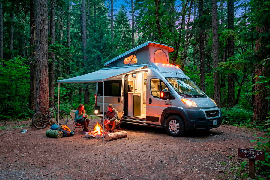 Cozy RV rental camping: A couple enjoys a campfire beside their camper van in a lush forest campsite at dusk.