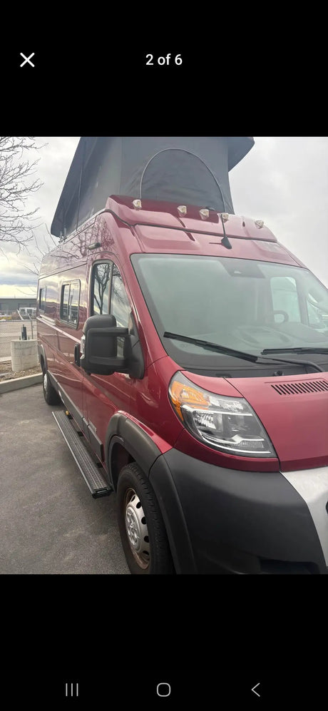 Red pop-top camper van for rv rental, parked, showing its front and side with running boards under an overcast sky.