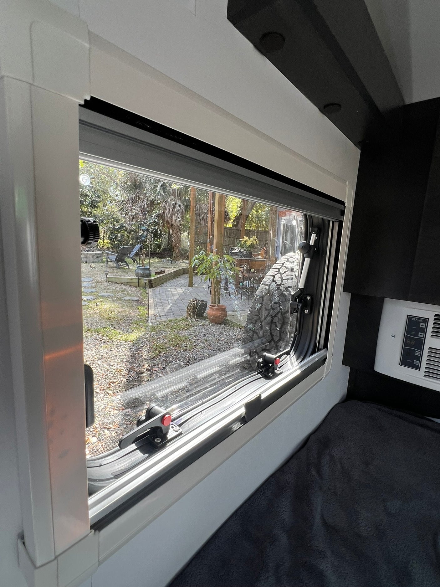 Bright RV rental interior looking out a window at a private backyard. Features include a view of a large tire, bed, and control panel.