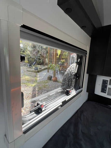 Bright RV rental interior looking out a window at a private backyard. Features include a view of a large tire, bed, and control panel.