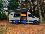 RV rental camper van in a forest with awning, lit interior, & chairs. Perfect for a cozy outdoor adventure.
