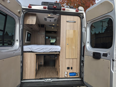 Rear view of an rv rental's interior, featuring a bed, under-bed storage, and a utility panel with hose, in a modern camper van.