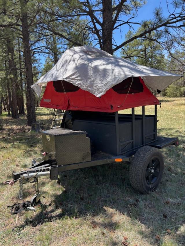 This camping trailer with a red rooftop tent & tarp is an excellent RV rental option for forest adventures.|This black off-road camping trailer with a tent frame is parked in a grassy forest. Great for your next RV rental adventure.|Red Yakima rooftop tent on a trailer — photo 1