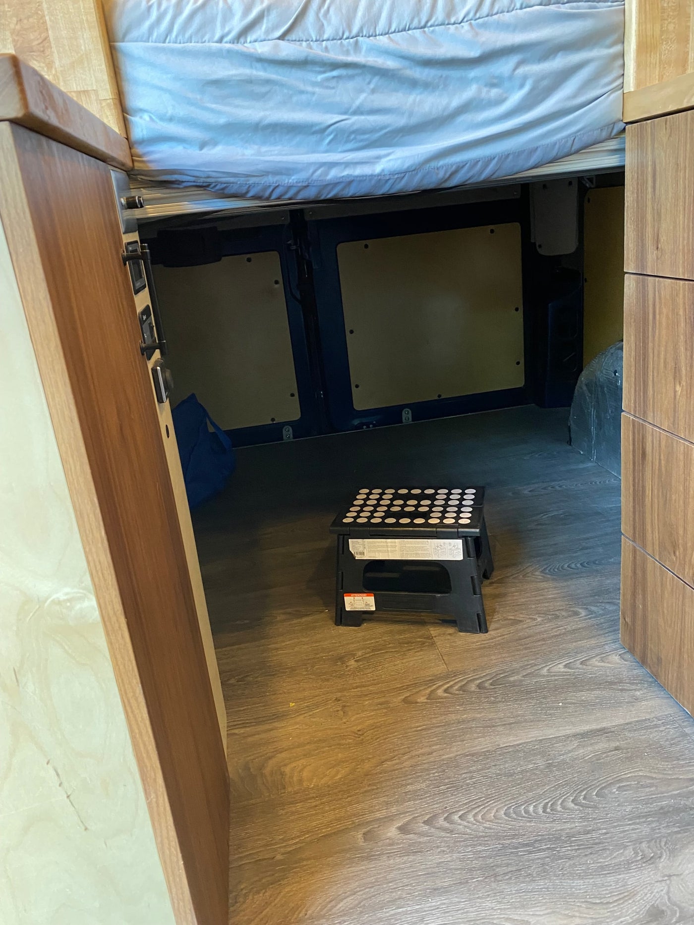 RV rental interior: A raised bed over under-bed storage, with a black step stool on wood-look flooring and wooden cabinets. — photo 10