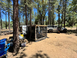 A white Jeep and an RV rental trailer with a side tent create a comfortable overlanding campsite in a sunny forest.