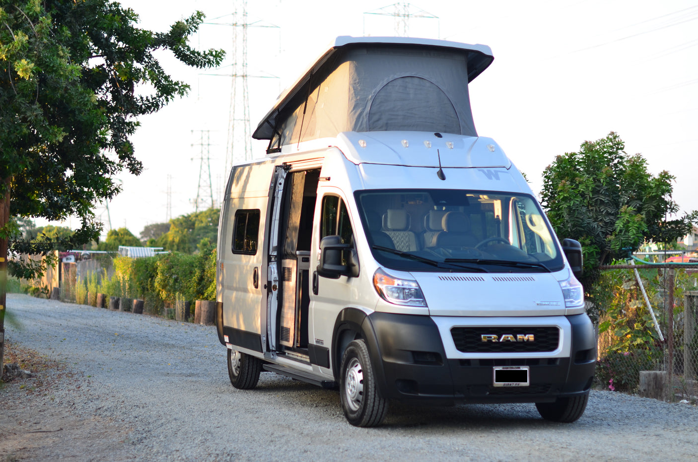 White Ram ProMaster camper van RV rental with pop-top roof on a gravel path, side door open, surrounded by trees.
