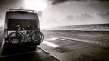 An RV rental, bikes on back, parked seaside on a paved path. Ocean waves and cloudy sky complete this beach adventure scene.