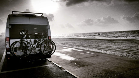 An RV rental, bikes on back, parked seaside on a paved path. Ocean waves and cloudy sky complete this beach adventure scene.